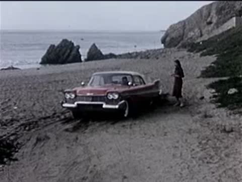 A red car parked on a sandy beach next to a woman standing nearby.
