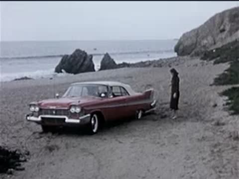 A red vintage car parked on a sandy coastal area next to a woman.