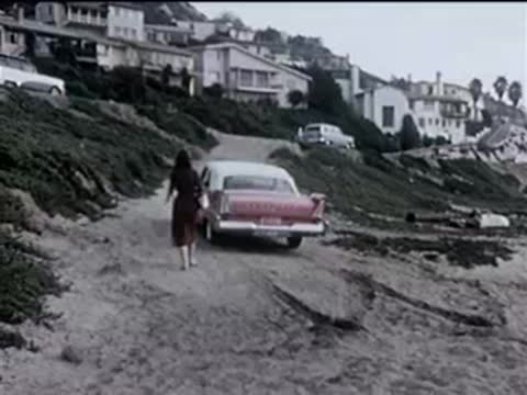 A woman walking away from a red car parked on a dirt road near houses.