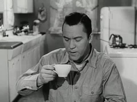 Man sitting in a kitchen drinking from a small white cup.