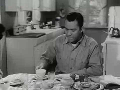 Man drinking coffee at a kitchen table with a woman nearby.