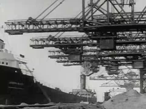 Large industrial cranes load materials at a harbor dock with ships.
