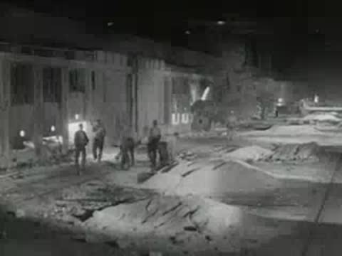 Workers handle raw materials inside large industrial factory setting, 1950s documentary footage.