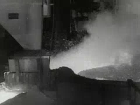 Workers converse near industrial equipment with smoke venting inside factory, 1950s.