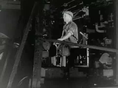 Operator sits at control console inside industrial facility, 1950s documentary footage.