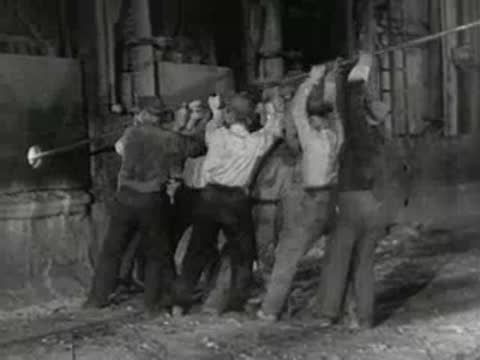 Group of workers uses long poles to work on industrial furnace, 1950s.