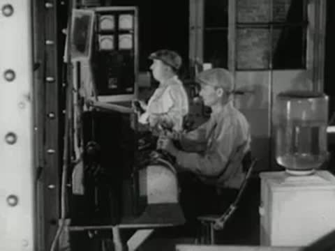 Two workers sit at control panels inside industrial facility, 1950s documentary.