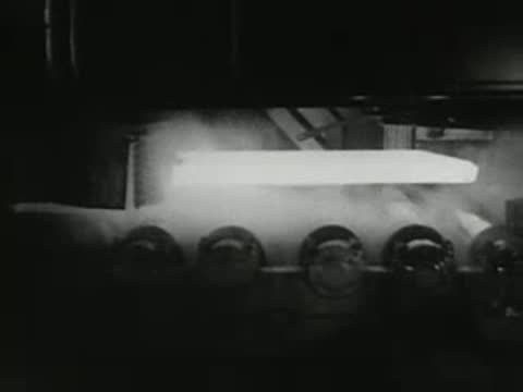 Workers oversee hot glowing metal slab moving along industrial rollers, 1950s.