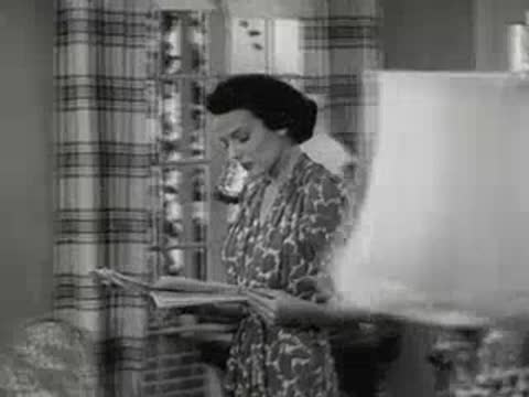 A woman in a kitchen looks at a newspaper near a window.