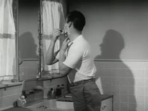 A man shaves his face with foam in a bright bathroom mirror.