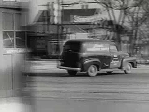delivery van driving away from hardware storefront on city street, 1950s