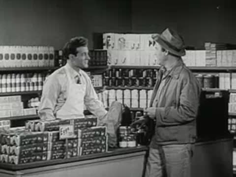 A grocery store clerk and a customer talking near shelves of goods