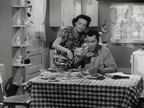 A woman walking up to a man eating at a table and serving him food