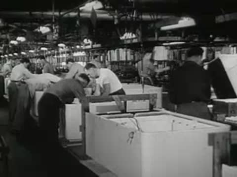 Line of workers assembling large white appliances in a factory.