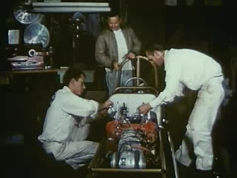 Two mechanics working on the engine of a dragster in a workshop.