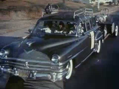 Vintage station wagon towing a dragster trailer and a man wearing sunglasses.