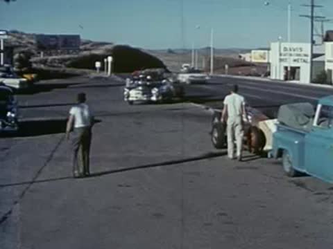 Men gather around a race car and truck at a roadside service station.
