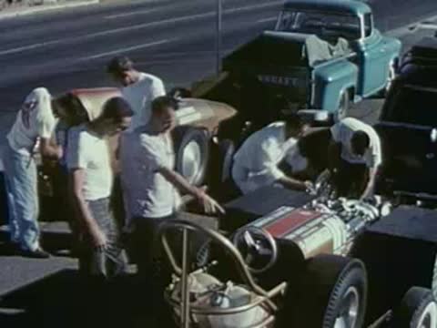 Mechanics work on the engine of a vintage race car outdoors.