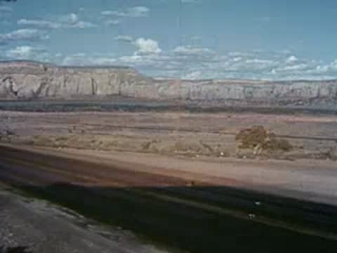 Cars drive along a desert highway near barren hills under a cloudy sky.
