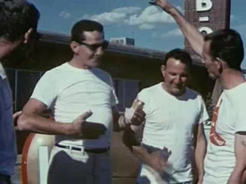 Three men converse outdoors near a vehicle with a fuel tank.