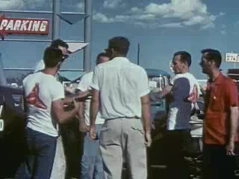 A group of men stand chatting near parked vintage cars outdoors.