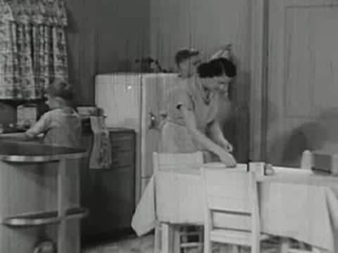 Boy standing by kitchen table while woman works nearby