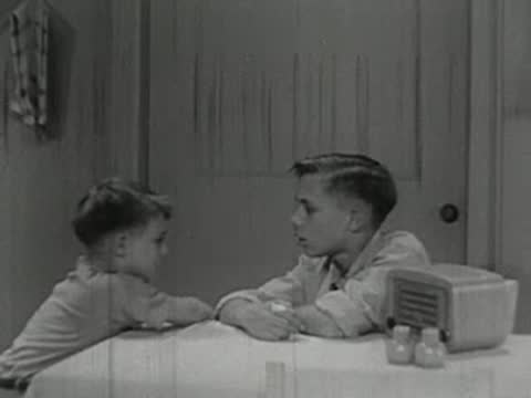 A young boy standing and gesturing to another boy sitting at a table.