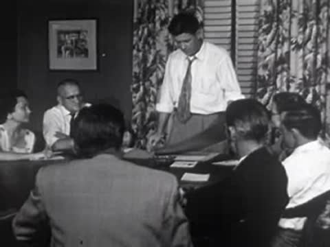 Man standing and presenting plans at a meeting table, 1950s office.