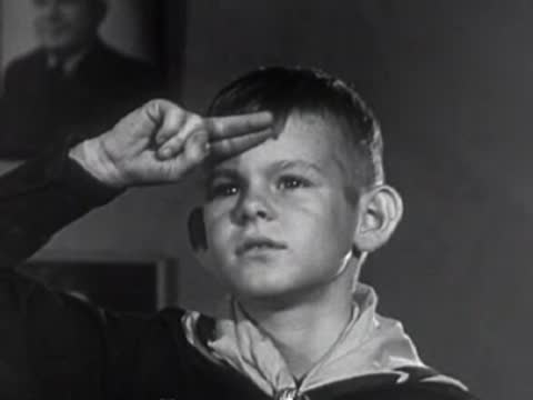 Cub scout saluting indoors, vintage uniforms, 1950s patriotic educational film scene.