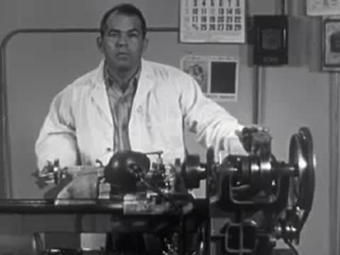 Man in lab coat operating lathe machine in a workshop, 1950s industrial scene.