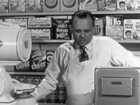 A grocery store clerk leaning over the counter, mid-century store shelves behind him.