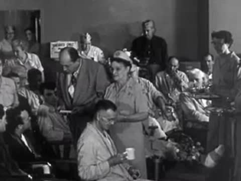 A crowded cafeteria or hospital ward where a woman serves coffee to seated patients.