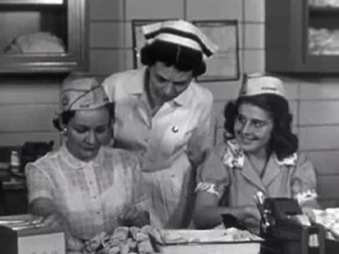 Two nurses in uniform working with papers at a desk, with a head nurse watching.