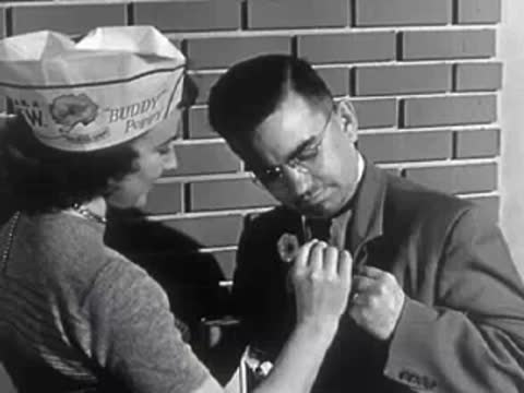 A woman pinning a Buddy Poppy flower onto a man's coat lapel near a brick wall.