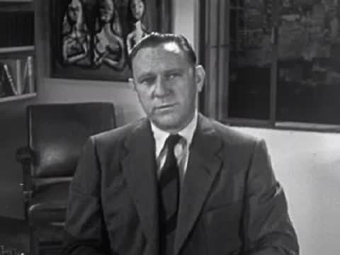 Man in suit talking directly to camera, 1950s style interview scene.