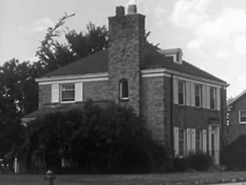 A two-story brick house with a stone chimney and large bushes in front.