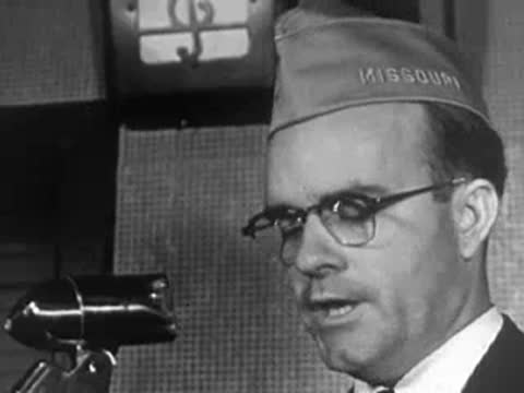 A man wearing glasses and a service cap speaking into a vintage microphone.