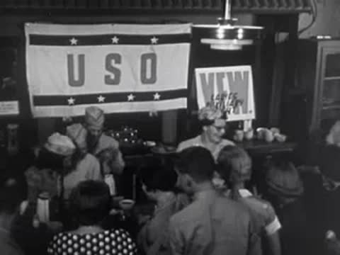 A group of people gathered in a USO hall with banners and decorations visible.
