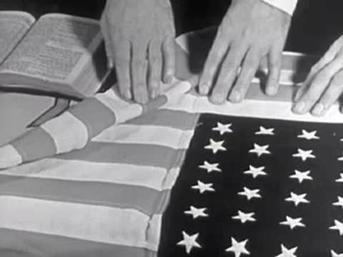 Close-up of hands folding an American flag next to a bible, 1950s.
