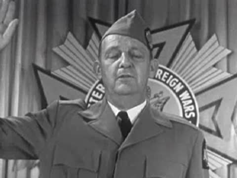 Men in uniform saluting with American flags and emblem in background, 1950s.