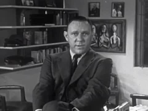 Man in suit sitting in an office speaking, 1950s.