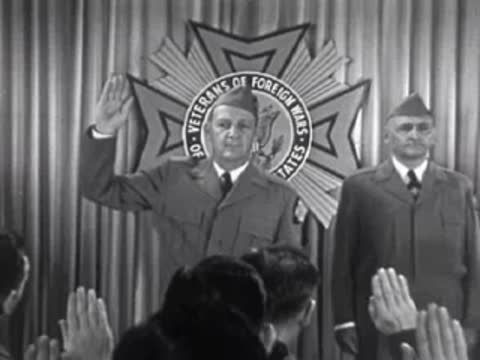 Men in uniform saluting in front of curtains and emblem, 1950s.