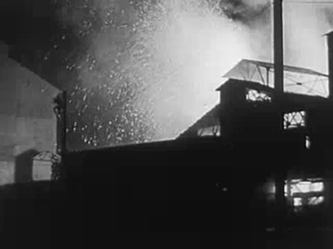 An industrial factory building at night with smoke billowing and bright lights illuminating the scene.