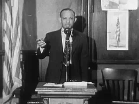 Black man in a suit speaking at a podium with microphone and flag