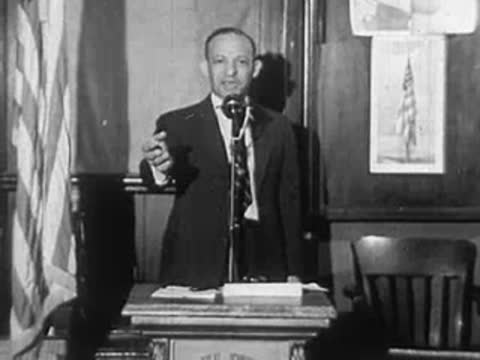 A man speaking at a podium into a microphone in front of flags.