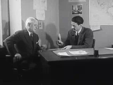 Two men sitting at a desk, shaking hands, office setting.