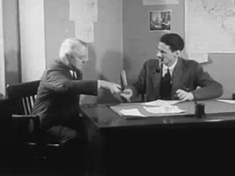 Two men at a desk talking and shaking hands, office background.
