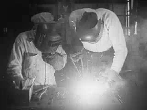 Two welders in protective gear working together on metal in a factory.
