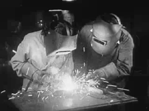 Workers welding metal parts at a workbench with sparks flying.