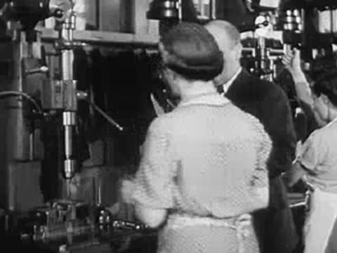 A woman operating a drill press while consulting with a man in a factory.